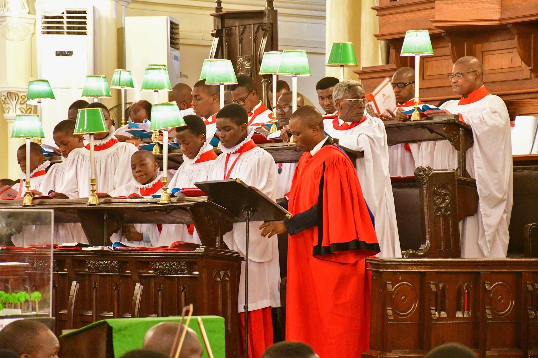 The choir of the Cathedral Church of Christ, Lagos, in red cassocks, white surplices and ruffs, singing in their stalls under green-shaded lamps, with the choirmaster in a red gown at the lectern.