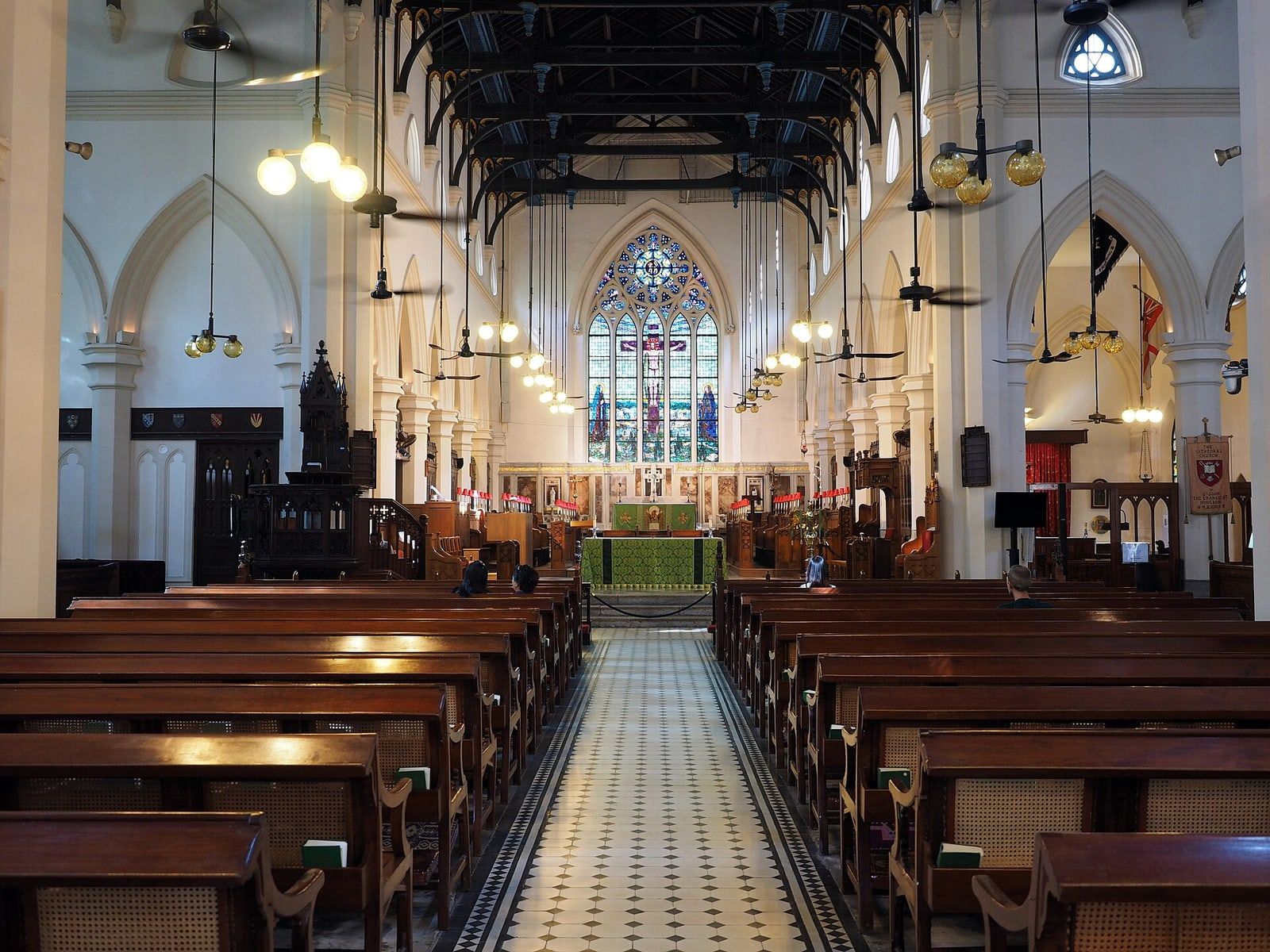 The white interior of St John’s Cathedral, Hong Kong: dark wooden pews, ceiling fans and a stained-glass east window.