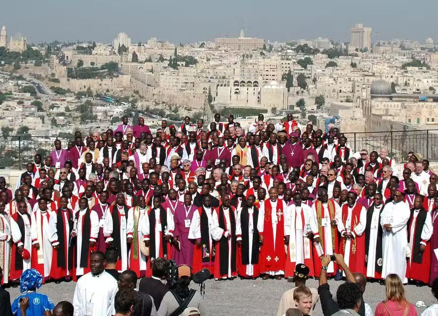 Hundreds of Anglican bishops and clergy in choir dress, many in red and white, gathered for a group photograph on a hillside with Jerusalem behind them.